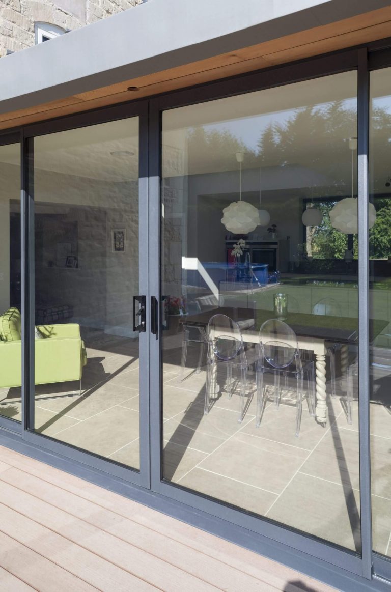 Grey-framed sliding glass patio doors leading into a modern dining and living space, with reflections of trees and sky visible on the glass.