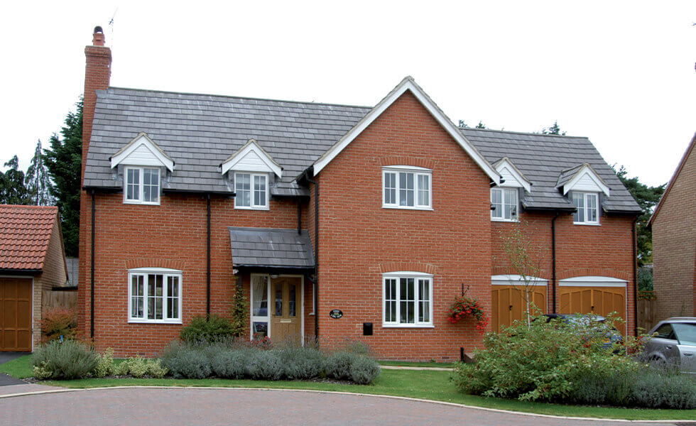 A two-story brick house with casement windows, featuring white frames and a central front door flanked by side windows. The home has a grey shingle roof with two dormer windows and a double garage with wooden doors. It is surrounded by a well-maintained garden with shrubs and a brick driveway, set against a suburban backdrop.