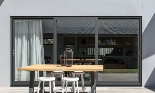 A modern patio with a wooden table and white stools in front of large sliding glass doors of a gray contemporary house, with a clear vase on the table.