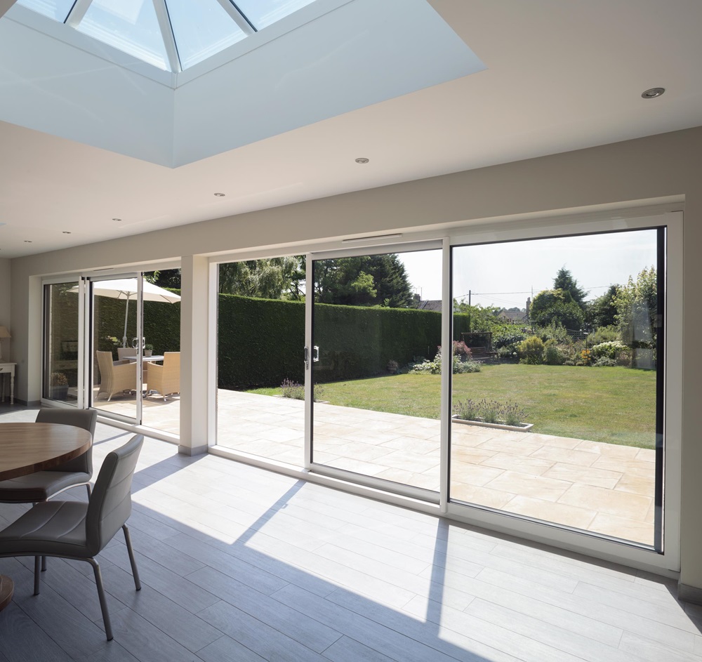 View from a modern dining room through large sliding glass doors opening to a patio and a beautifully landscaped garden, with abundant natural light streaming from a skylight overhead.